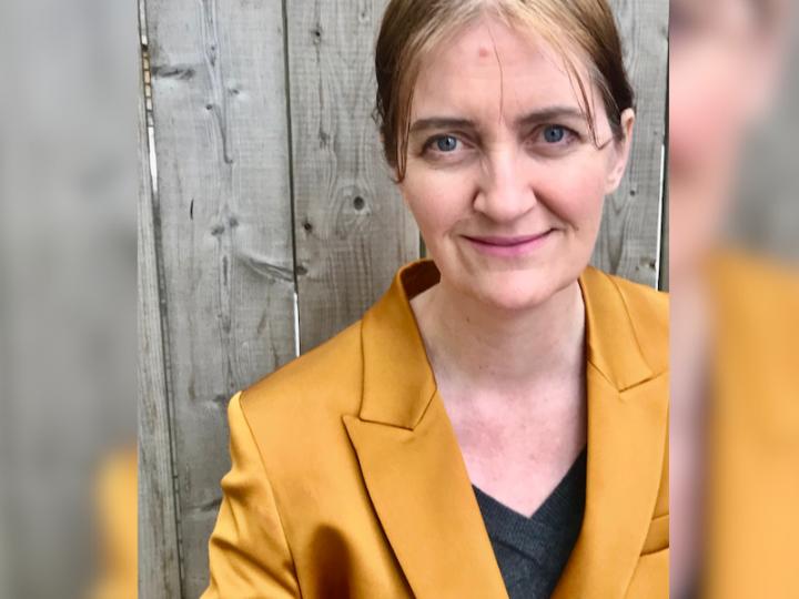 Author Emma Donoghue wearing a metallic gold blazer and black V-neck tee. Her light blond hair is pulled back behind her ears. She is in front of a grey fence.