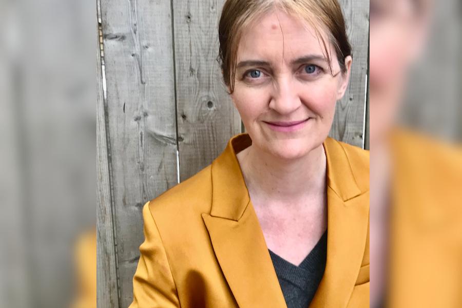 Author Emma Donoghue wearing a metallic gold blazer and black V-neck tee. Her light blond hair is pulled back behind her ears. She is in front of a grey fence.