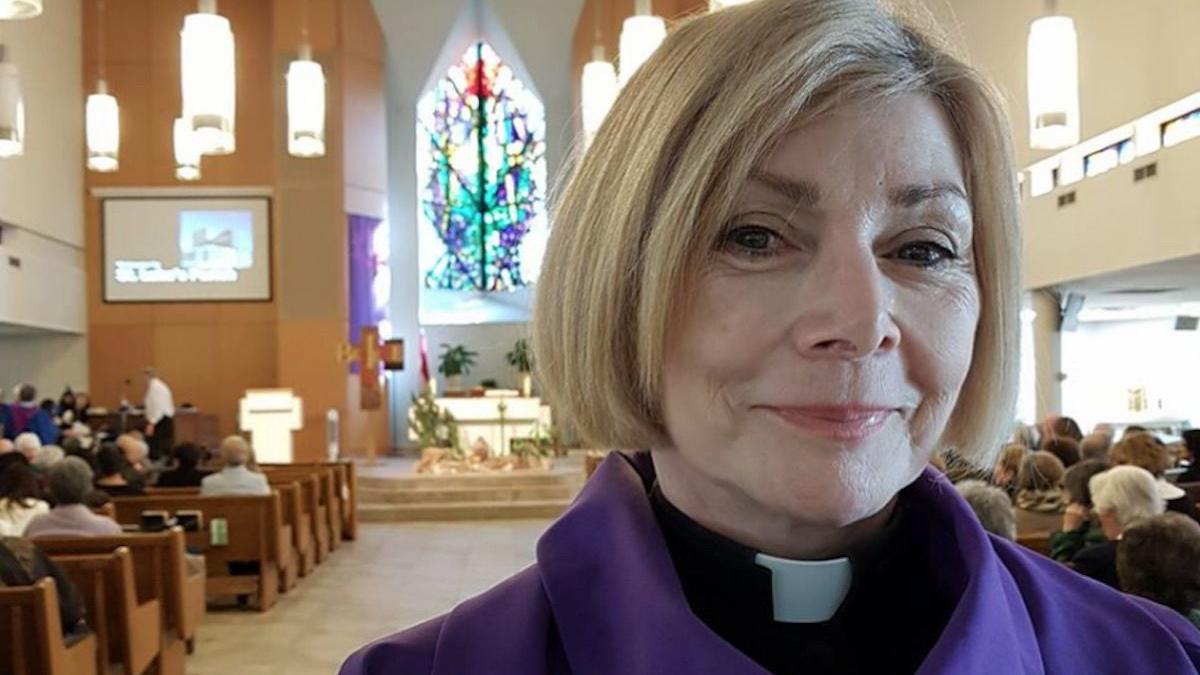 Rev. Cheri DiNovo standing inside a church with congregation members sitting behind her, looking at a screen.