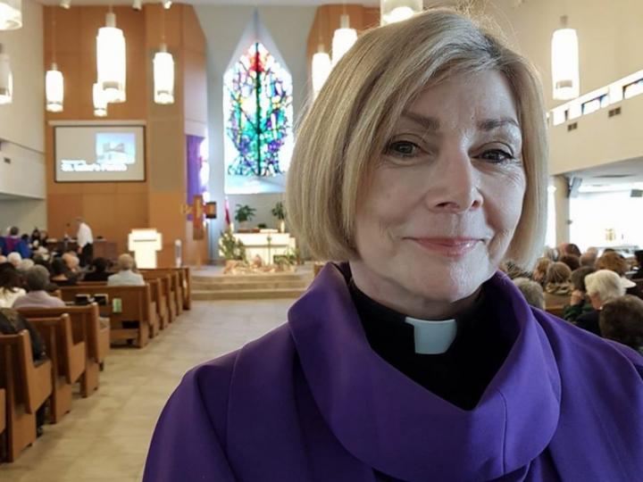Rev. Cheri DiNovo standing inside a church with congregation members sitting behind her, looking at a screen.