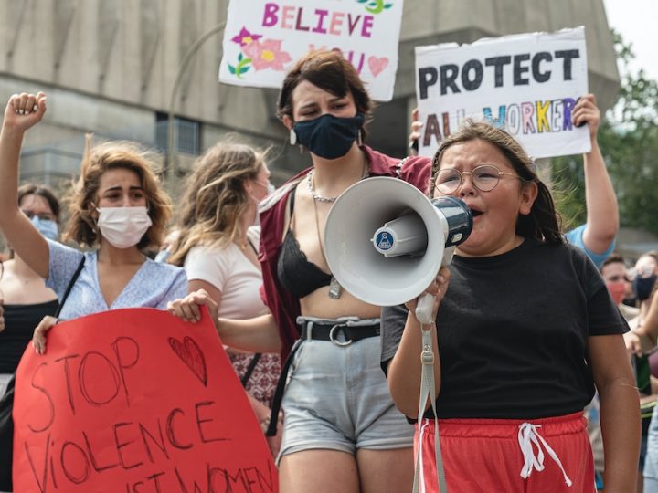 Women at a protest standing behind a young girl with tears in her eyes as she screams into megaphone. The girl is dressed in a black t-shirt and red shorts.