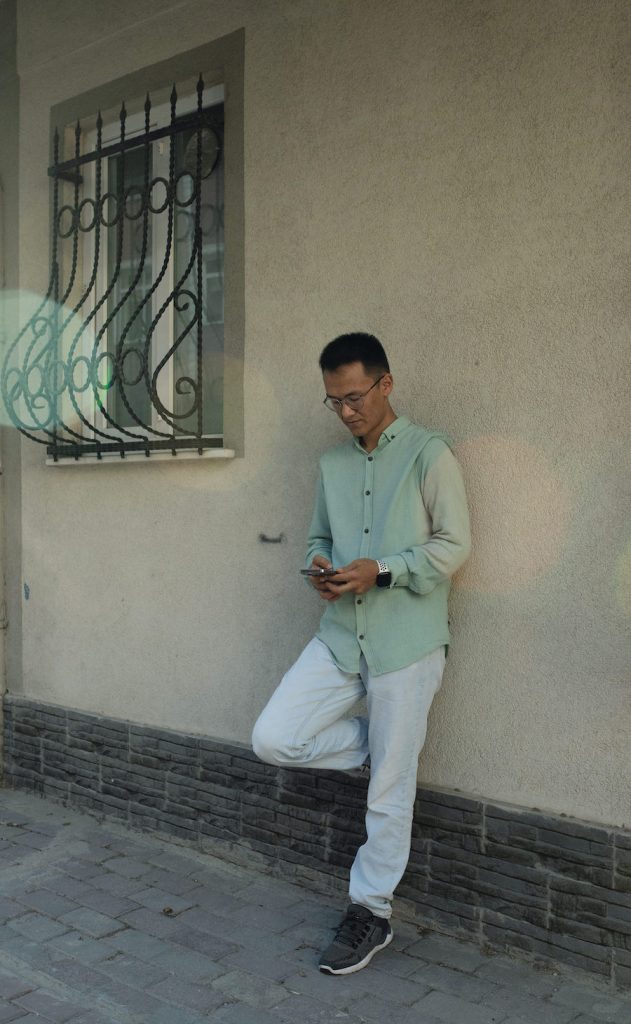 Man in a green button shirt and white pants standing against a white wall.