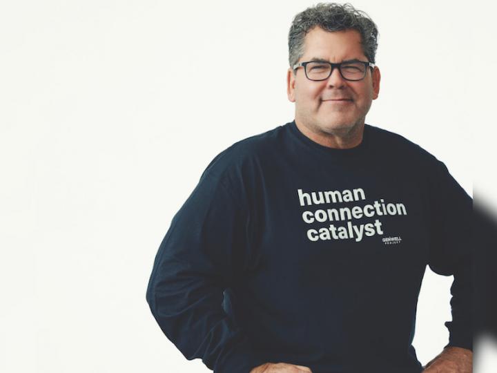 Man sitting on a stool in a navy blue long sleeve that reads: human connection catlyst. He is wearing dark blue jeans and black frame glasses.