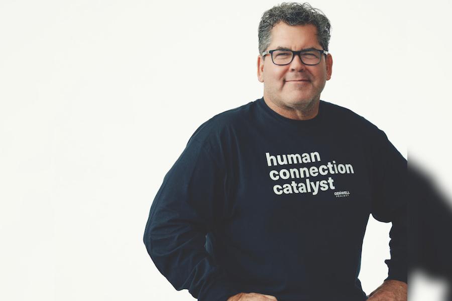 Man sitting on a stool in a navy blue long sleeve that reads: human connection catlyst. He is wearing dark blue jeans and black frame glasses.
