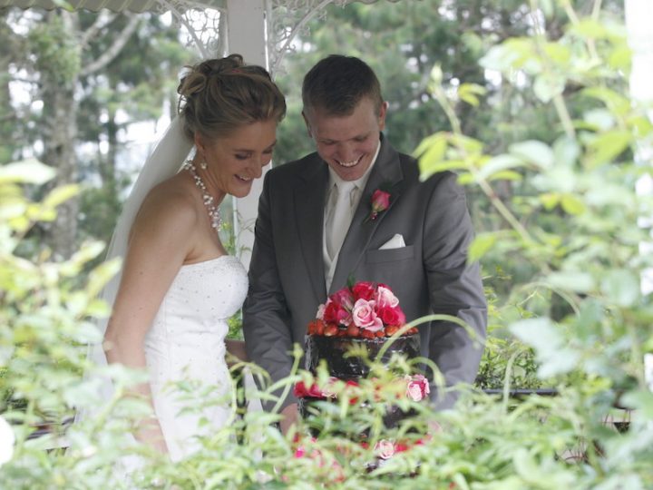 Bride and groom dressed in a white dress and a grey suit, cutting a chocolate cake with red and pink flowers.