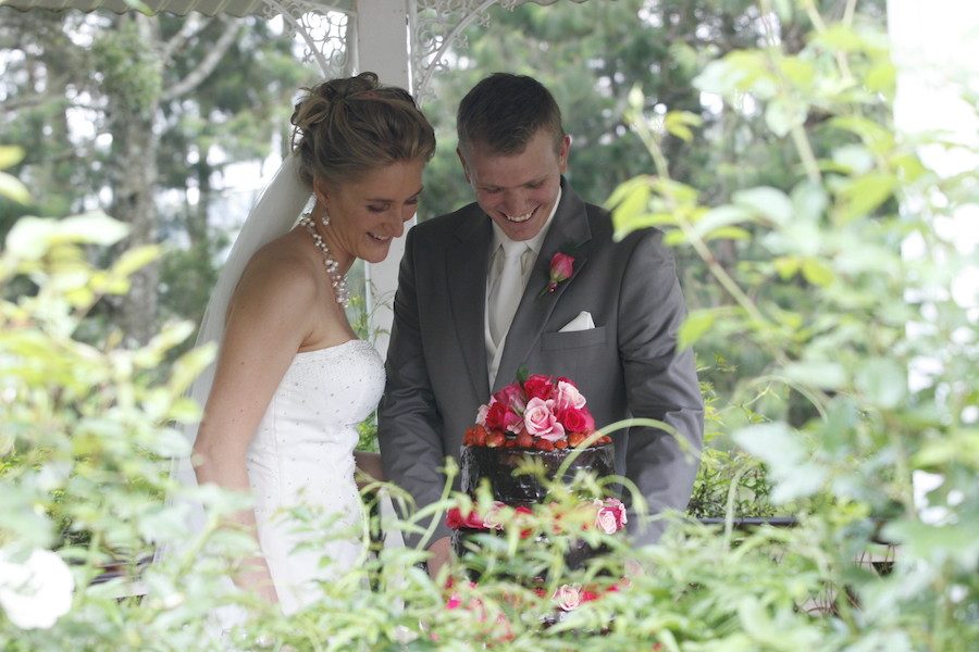 Bride and groom dressed in a white dress and a grey suit, cutting a chocolate cake with red and pink flowers.