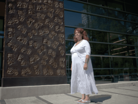 White woman in white dress stands before wall of hand impressions