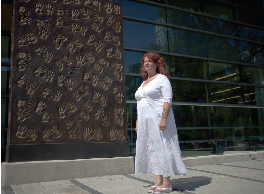 White woman in white dress stands before wall of hand impressions