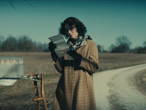 Woman with short brown hair dressed in a brown plaid trench coat, multi-coloured scarf and black gloves holding a newspaper.