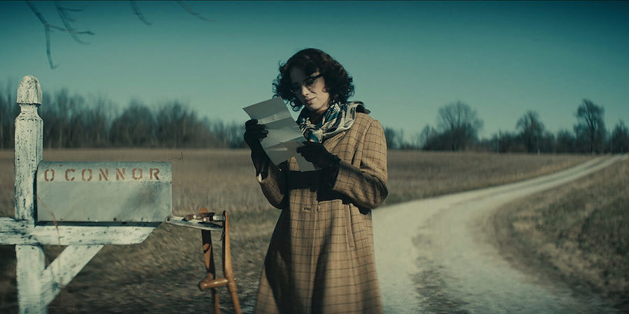 Woman with short brown hair dressed in a brown plaid trench coat, multi-coloured scarf and black gloves holding a newspaper.