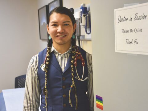 Person with two long black braids wearing a navy blue vest over a striped black and white shirt. They are standing beside a grey door with a pride flag and a sign that reads "Doctor in Session. Please be Quiet. Thank You"