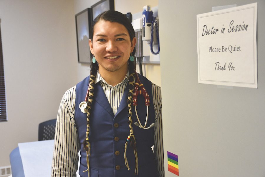 Person with two long black braids wearing a navy blue vest over a striped black and white shirt. They are standing beside a grey door with a pride flag and a sign that reads "Doctor in Session. Please be Quiet. Thank You"