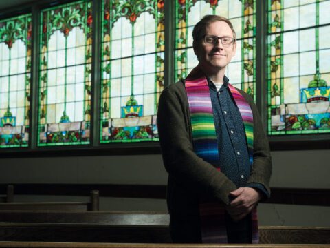 Man wearing a dark blue dress shirt, green cardigan, and multi-coloured scarf around his neck. He has short blond cropped hair and glasses. He is standing in front of dark brown pews and stained glass windows of green, blue, yellow, orange and red.