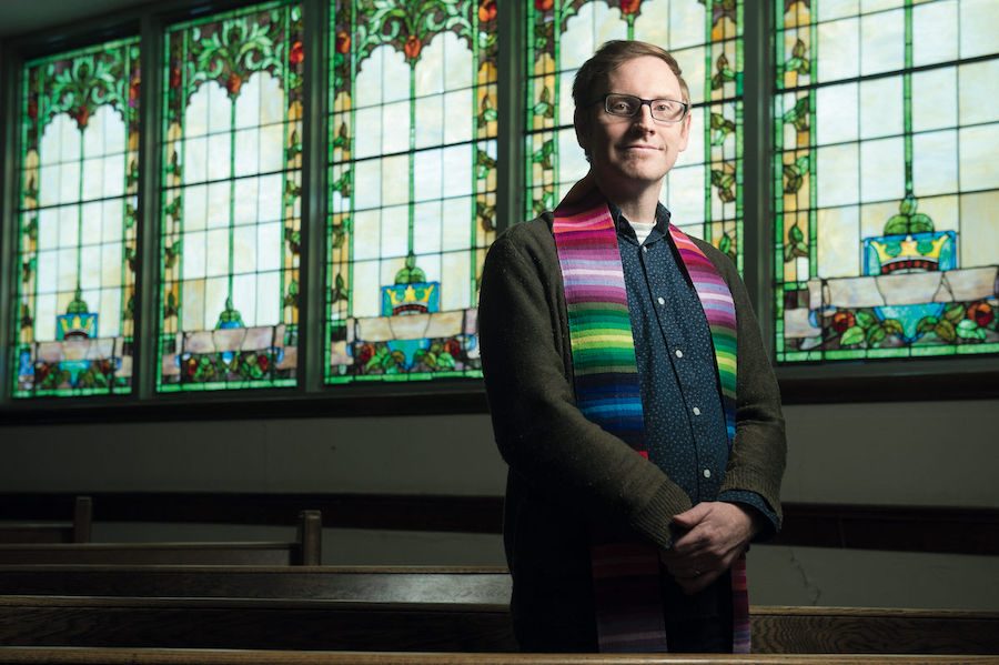 Man wearing a dark blue dress shirt, green cardigan, and multi-coloured scarf around his neck. He has short blond cropped hair and glasses. He is standing in front of dark brown pews and stained glass windows of green, blue, yellow, orange and red.