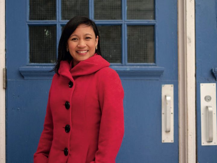 Woman with shoulder-length black hair standing in a red coat in front of a blue door.