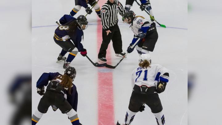Women hockey players competing on ice with a referee in the middle.