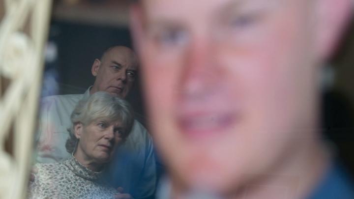 Older white couple in front of blurry large image of young man