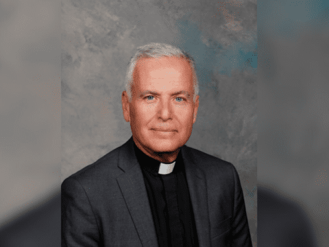 A person wearing black top, dark grey jacket and a white priest collar in front of a grey background.
