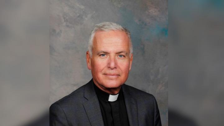 A person wearing black top, dark grey jacket and a white priest collar in front of a grey background.