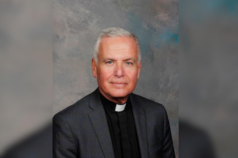 A person wearing black top, dark grey jacket and a white priest collar in front of a grey background.