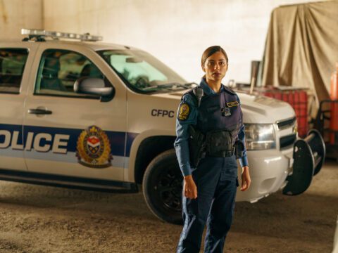 A woman with brown skin and dark brown hair with light brown highlights dressed in a navy blue police uniform stands in front of a white and blue police car.
