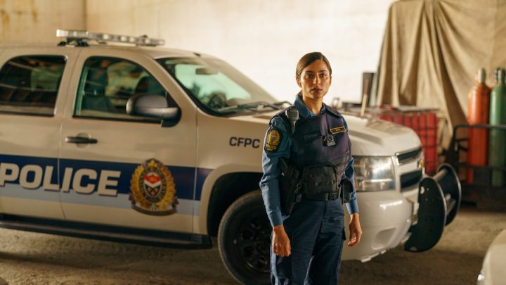 A woman with brown skin and dark brown hair with light brown highlights dressed in a navy blue police uniform stands in front of a white and blue police car.