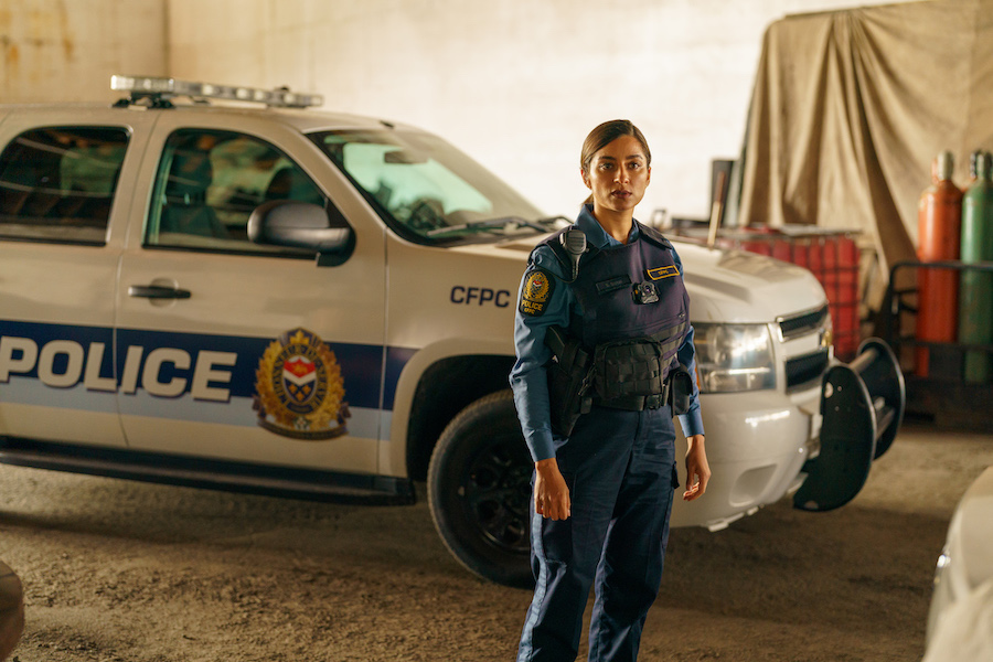 A woman with brown skin and dark brown hair with light brown highlights dressed in a navy blue police uniform stands in front of a white and blue police car.