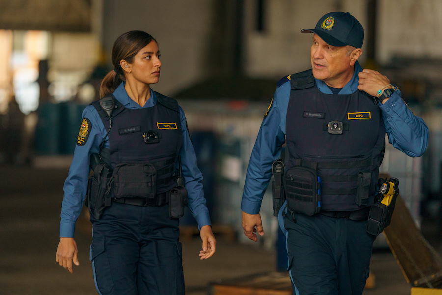 A woman with brown skin and dark brown hair with light brown highlights dressed in a navy blue police uniform stands next to a man with white skin and a police cap dressed in a navy blue police uniform.
