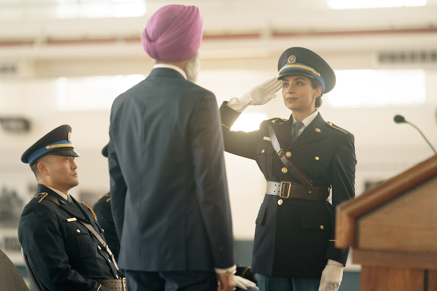 A woman in a navy blue police officer uniform salutes a man in a dark grey suit and light purple turban. There is a brown podium to their right and a man dressed in a police uniform sitting on a chair to their left.