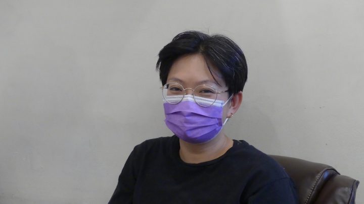 Person with short black hair is wearing a purple medical mask and a black t-shirt, sitting in a dark brown chair.