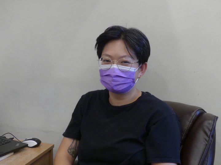 Person with short black hair is wearing a purple medical mask and a black t-shirt, sitting in a dark brown chair.