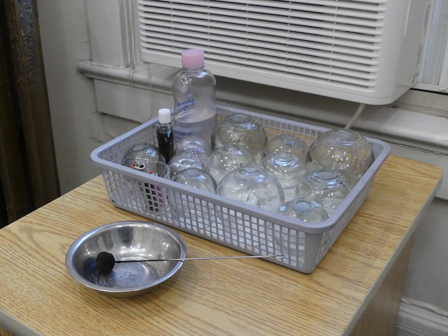 Plastic grey container with glass cups and body oil resting on a wooden table. There is a silver steel bowl in front of it with a metal rod inside it. 