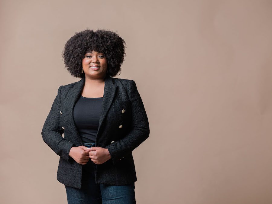 Black woman with black afros dressed in a black blazer, black top and dark blue jeans. She is smiling with her hands clasped in front of her stomach.