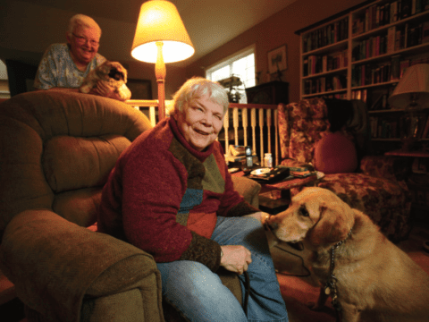 Older white woman sitting in a dimly lit living room. She is petting large dog. The woman looks happy and inquisitive but the dog looks bored. There is another older woman in the background smiling, holding a tiny dog, who is hopefully not as bored.