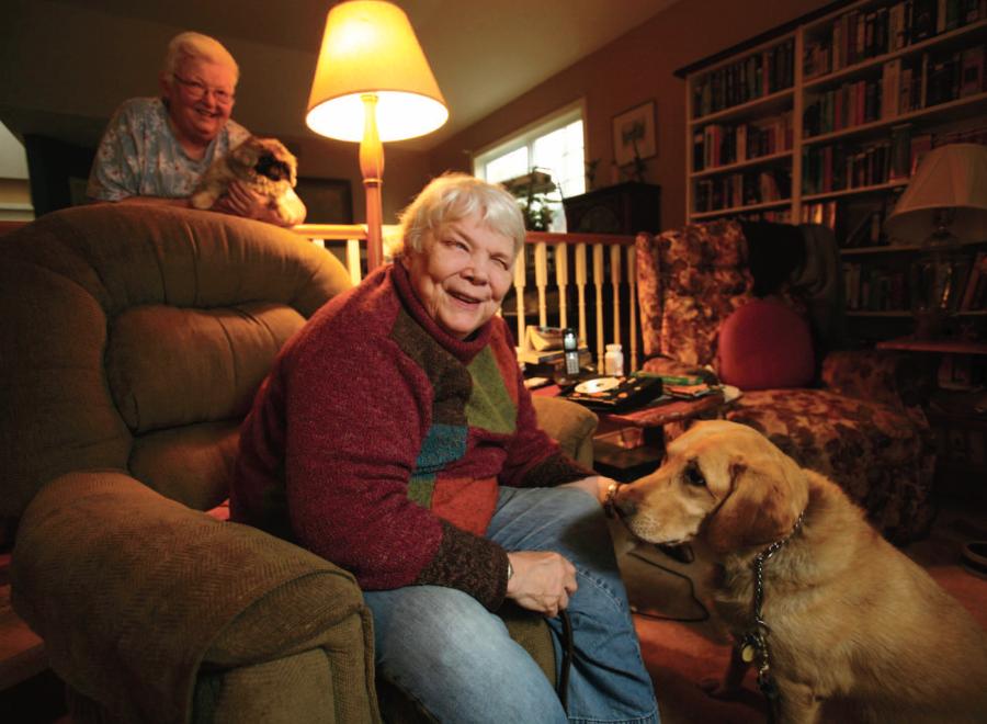 Older white woman sitting in a dimly lit living room. She is petting large dog. The woman looks happy and inquisitive but the dog looks bored. There is another older woman in the background smiling, holding a tiny dog, who is hopefully not as bored.