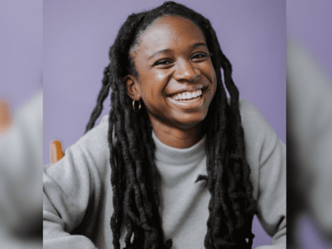 Black woman with Black locs smiling. She is wearing a grey sweater, seated in a wooden chair with a purple background.
