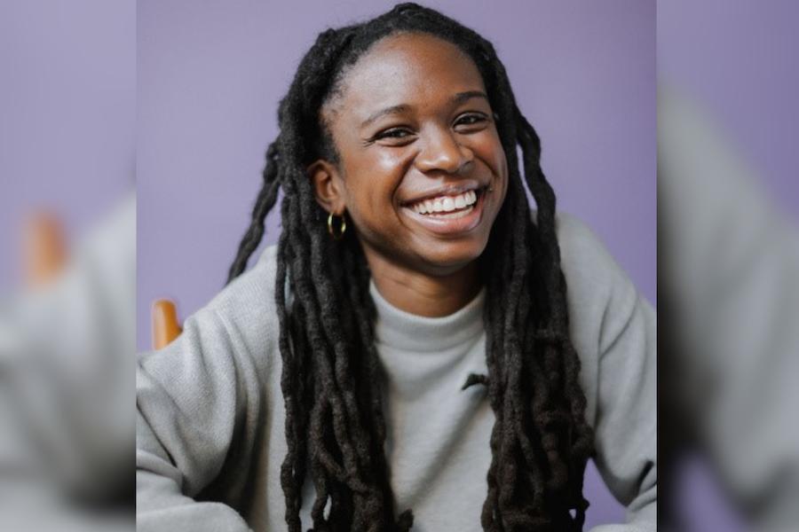 Black woman with Black locs smiling. She is wearing a grey sweater, seated in a wooden chair with a purple background.
