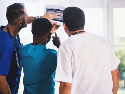 Three male doctors reviewing x-ray results. One has an olive skin tone wearing royal blue scrubs and a stethoscope, one has a black skin tone wearing teal scrubs and one has a fair skin tone wearing white scrubs.