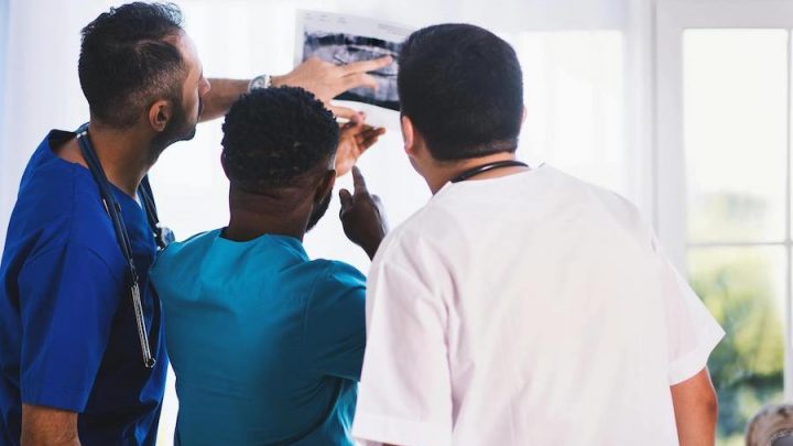 Three male doctors reviewing x-ray results. One has an olive skin tone wearing royal blue scrubs and a stethoscope, one has a black skin tone wearing teal scrubs and one has a fair skin tone wearing white scrubs.