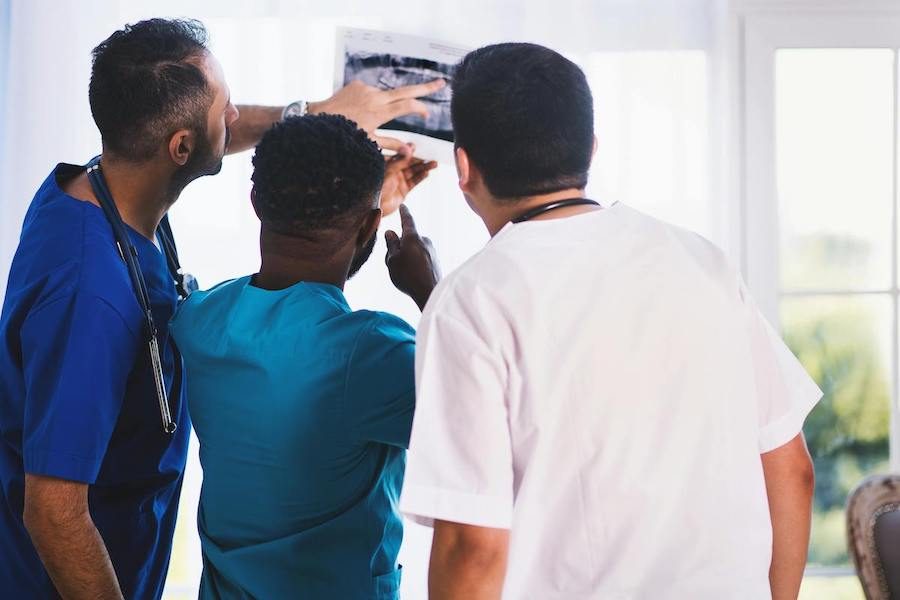 Three male doctors reviewing x-ray results. One has an olive skin tone wearing royal blue scrubs and a stethoscope, one has a black skin tone wearing teal scrubs and one has a fair skin tone wearing white scrubs.