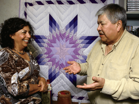 Indigenous woman and man in front of quilt