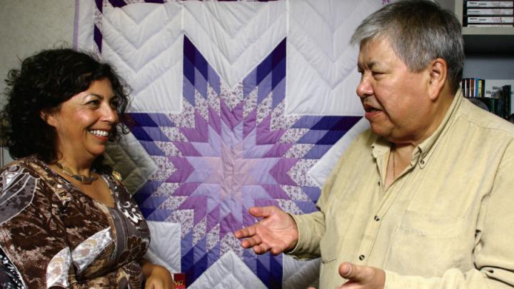 Indigenous woman and man in front of quilt