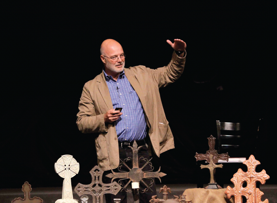 Bald-headed white man with grey beard in sports jacket standing in front of sacred items