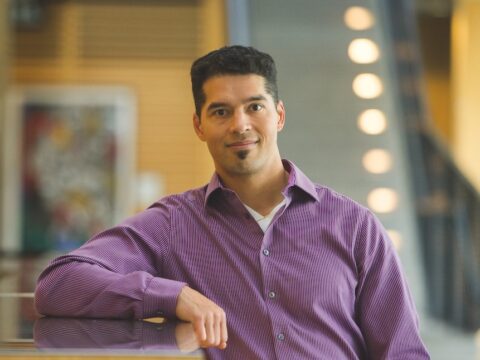 A man with olive skin, black hair and a goatee wearing a purple button-up shirt stands with his right arm resting on a counter.