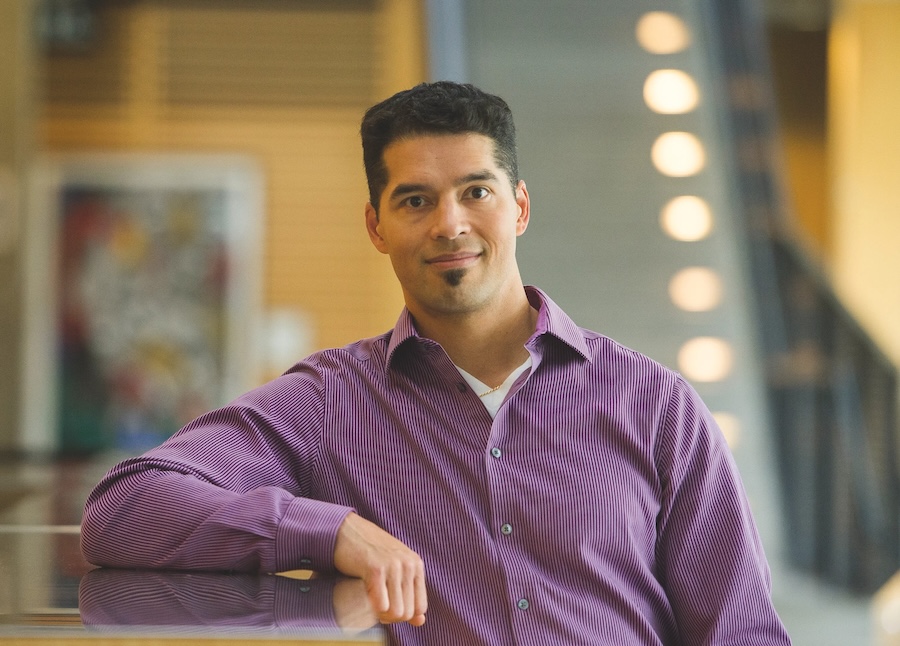 A man with olive skin, black hair and a goatee wearing a purple button-up shirt stands with his right arm resting on a counter.