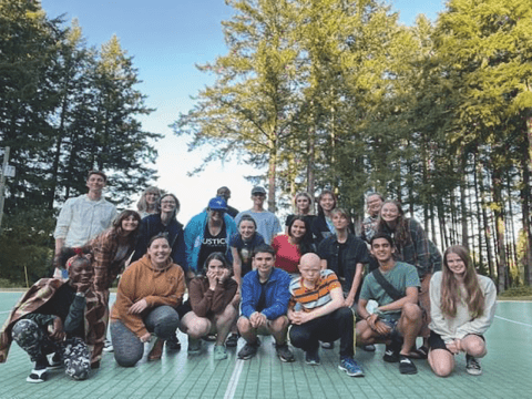 A group of youths from different backgrounds and races are standing in front of trees and blue skies. The youth in the front row are kneeling down.