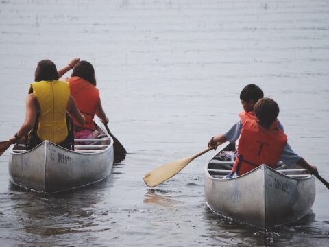 Two canoes with two children in each are paddling in a lake. The children are wearing yellow and orange vests and the perspective of the image is looking from behind so you see their backs. They are holding paddles in their hands.