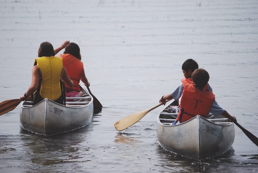 Two canoes with two children in each are paddling in a lake. The children are wearing yellow and orange vests and the perspective of the image is looking from behind so you see their backs. They are holding paddles in their hands.