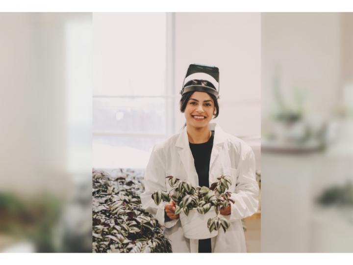 A brown-skinned woman with black har wearing safety glasses and a white lab coat, black shirt, is smiling and posing. She is holding a pothos plant in her hands. The plant is in a white pot with white and green leaves.
