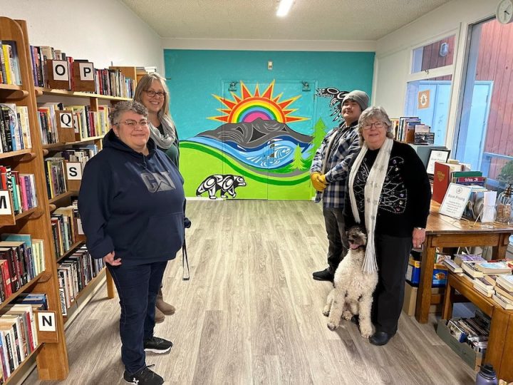 Four people stand in front of a colourful mural in a room full of books and shelves. The mural features a blue sky with a red sun and a rainbow in the sun. The sun is rising above grey mountains and blue rivers. The rivers have salmon painted in it and runs along a green landscape and pine trees. In the sky there is a black eagle painted in an Indigenous style. On the green land, there is a black and grey bear painted in an Indigenous style. The four people standing in front of the mural include three women and one young man. One woman is the artist. She is wearing a black hoodie and has short curly black and grey hair. She is wearing glasses. Behind the artist is a taller white woman with blond hair and is wearing a white scarf and black shirt. On the other side, in the front is a middle-aged woman with white hair, white skin in a black shirt and pants with a long white scarf that has silver stars on it. She is holding the head of a black and white dog with curly fur who is sitting in front of her. Behind this woman is a young man with brown skin. He is wearing a blue and white plaid shirt, yellow gloves, black pants and a grey toque. He is smiling.